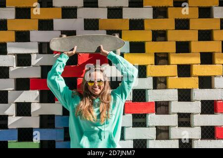 Young woman lifting skateboard while standing against brick wall on sunny day Stock Photo