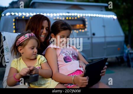 Smiling mother with daughters using digital tablet while sitting against motor home at dusk Stock Photo