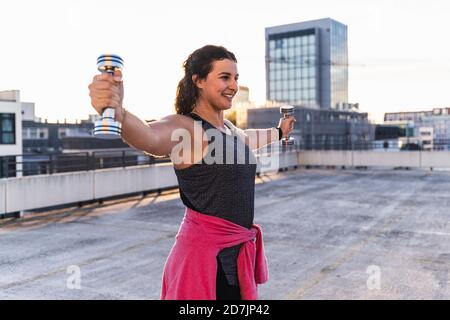 Smiling young woman with arms outstretched lifting dumbbells on terrace at sunset Stock Photo