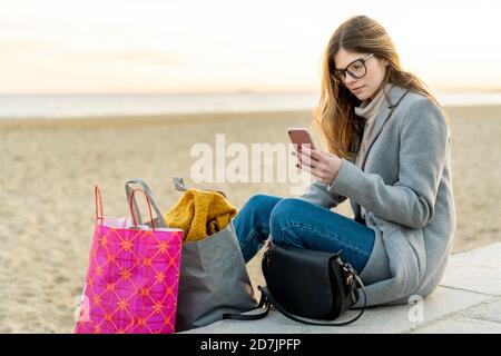 Beautiful young woman using smart phone while sitting with shopping bags at beach during sunset Stock Photo