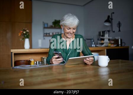 Senior woman listening to music while using smart phone and digital tablet at home Stock Photo