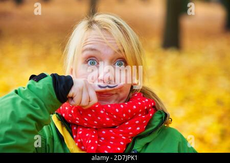 funny portrait of young woman in park in autumn with mustache painted on her finger Stock Photo