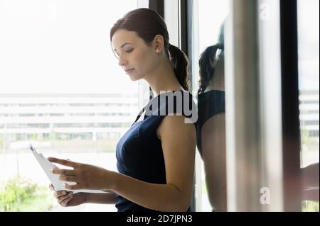 Businesswoman reading documents while standing by window in office Stock Photo