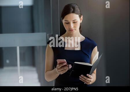 Confident businesswoman holding diary using smart phone while standing against wall Stock Photo