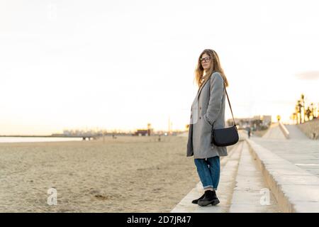 Thoughtful young woman with hand in pocket standing on promenade at beach while looking away during sunset Stock Photo