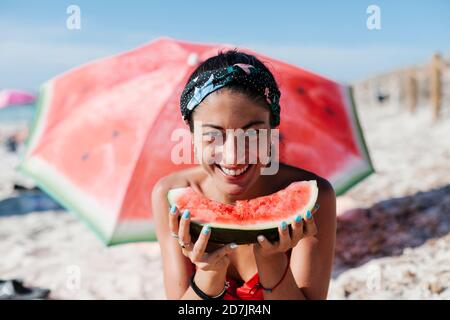 Close-up of smiling woman holding watermelon while sitting against red umbrella at beach Stock Photo