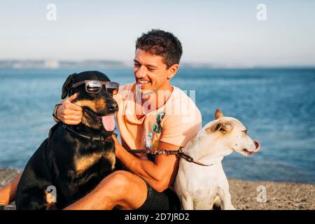 Smiling mid adult man playing with dogs while sitting at beach against sky Stock Photo