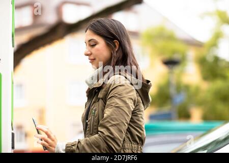 Beautiful woman using smart phone for renting electric car at charging station Stock Photo