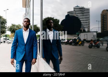 Confident male African entrepreneur walking by reflection of himself on window in city Stock Photo