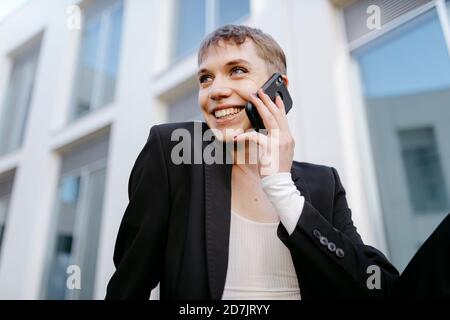 Close-up of smiling trans young man talking over mobile phone while sitting against building Stock Photo