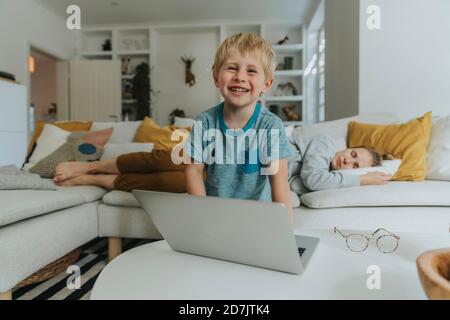 Smiling boy using laptop with mother sleeping in background at home Stock Photo