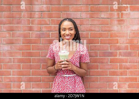 Young woman drinking smoothie while standing against brick wall Stock Photo