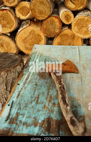 Old rusty ax lies on the blue shabby table near logs of birch wo Stock Photo