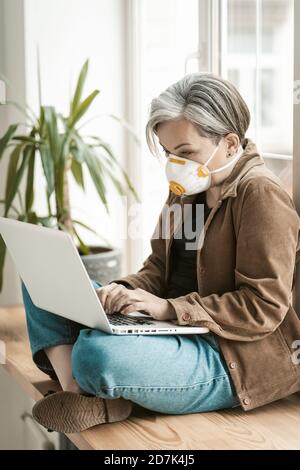 Greying businesswoman works laptop from home. Aged woman wearing protective mask uses mobile internet on computer sitting at windowsill. Qurarantine Stock Photo