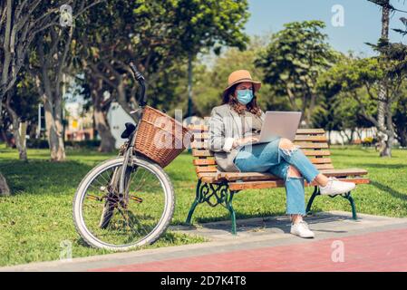 Woman works sitting in a park and protects herself with a medical mask. Portrait of a fashionable woman who enjoys the outdoors and writes on her blog Stock Photo