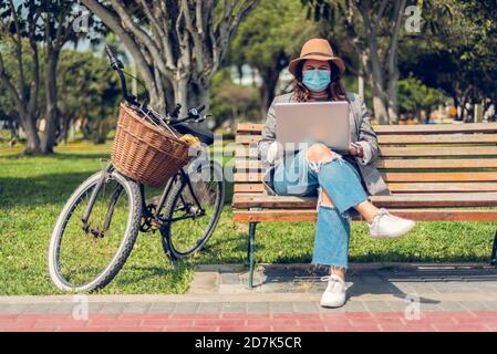 Woman works sitting in a park and protects herself with a medical mask. Portrait of a fashionable woman who enjoys the outdoors and writes on her blog Stock Photo