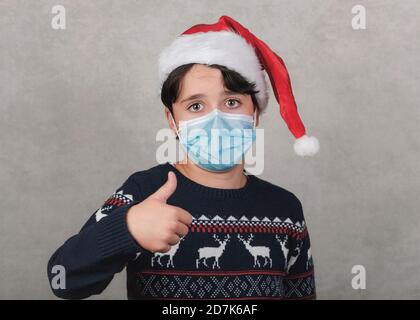 Merry Christmas, kid with medical mask wearing Santa Claus hat showing thumb up  over gray background Stock Photo