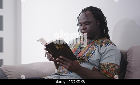 African Man in traditional dress with rosary reading the Holy Bible sitting on the sofa. High quality photo Stock Photo