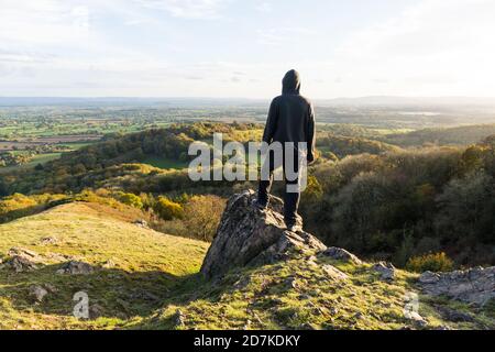 A hooded man, standing on a rocky outcrop on top of a hill, Looking out across the countryside in October. Malvern Hills, UK Stock Photo