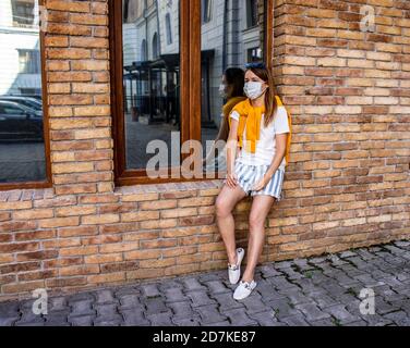 Young Caucasian woman in a protective mask in a white T-shirt and a yellow sweatshirt on a city street at the window of a brick building. Protection f Stock Photo