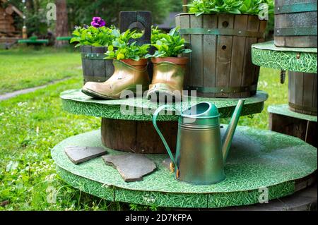 Hand made flower pot from old rubber boots and wooden flower pots and a metal watering can in the summer garden. Eco concept, eco design Stock Photo