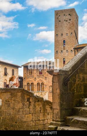Tourist woman making pictures with her smartphone from the stairs of the Duomo of San Gimignano, with Torre Chigi in the background, Tuscany, Italy Stock Photo