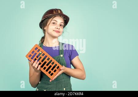 building her future house. happy childhood. kid wear protective helmet. protect head on construction site. teen girl hold brick. little builder with brick. Stock Photo