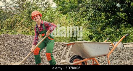 girl with wheelbarrow of rubble. kid working on construction site. teen girl takes out rubble from wheelbarrow. kid with shovel loading crushed stones. Laying the foundation, building project. Stock Photo