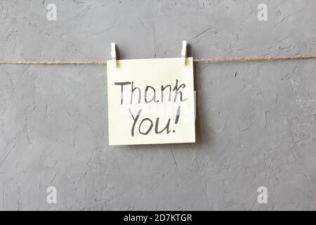 Note Thank you! is written on a yellow sticker that hangs with a clothespins on a grey background Stock Photo