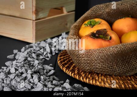 sack of delicious persimmons on a wicker plate, a vintage wooden box and stones, in shades of orange, brown and gray Stock Photo