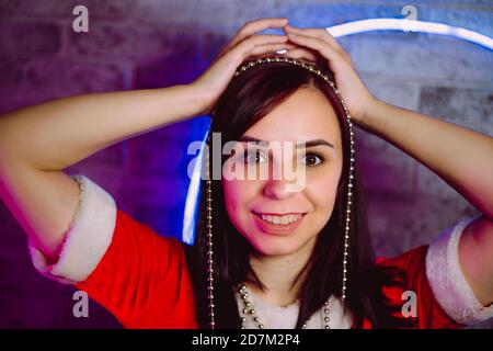 Portrait of young woman in Santa Claus suit with beads against illuminated wall. Close up of pretty female in Christmas costume holding beads. Concept Stock Photo