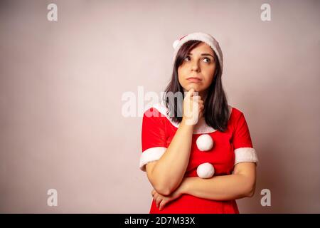 Young thoughtful woman in Santa Claus suit propping up her chin on grey background. Close up of female in Christmas hat pondering about gifts. Concept Stock Photo