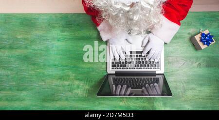 Aerial shot of Santa Claus working with his hands on a laptop keyboard together with a small gift on a green wooden desk, you can see a detail of his Stock Photo