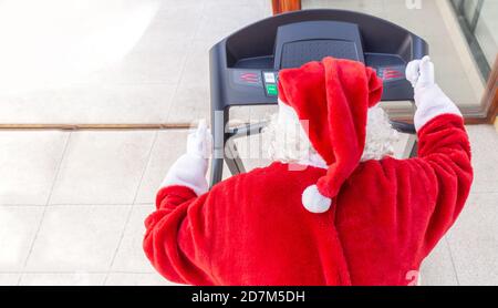 Santa Claus running on a treadmill in a gym at Christmas, shot from the back where you can see the white hair and the hat Stock Photo