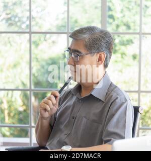 An Asian middle-aged man holding a pen thinking about solving problems on a desk in an office office. Stock Photo