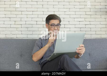 An Asian middle-aged man holding a notebook in his hand is sitting on a sofa taking a video call. Stock Photo