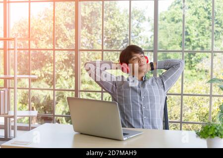 A young Asian businessman, listening to music on headphones on a desk on a balcony window. Stock Photo