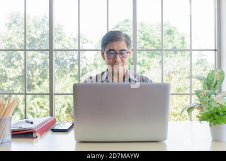 An elderly Asian man is learning how to trade online with a laptop on his desk. Stock Photo