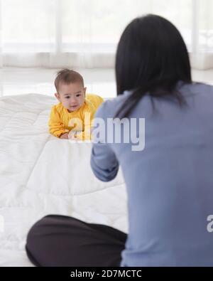 An Asian infant who is practicing crawling With her mother sitting on the other side of her soft white cloth in the bedroom. Stock Photo