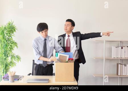 An Asian employee was fired from his job and was packing his personal belongings in paper boxes with a manager standing by him. Stock Photo