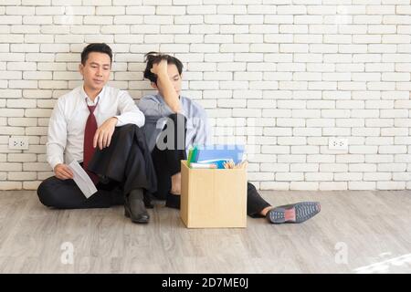 Two male employees sat on the floor talking in front of each other with a cardboard box for personal use after being fired from work. Stock Photo