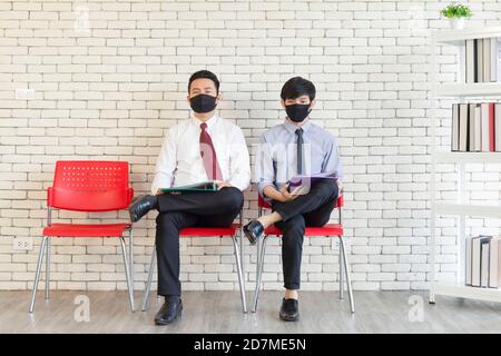 Two Asian men sit in clear red plastic chairs Matt gag to prevent COVID-19 waiting for a job interview. Stock Photo