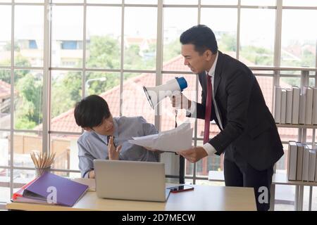An angry Asian man-manager shouting in a loudspeaker in his subordinate's ear to grab documents. Stock Photo