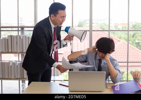 An angry Asian man-manager shouting in a loudspeaker in his subordinate's ear to grab documents. Stock Photo