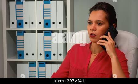 Woman speaking on phone during work time in modern office. Hispanic entrepreneur working in professional workplace in personal corporate company talking on smartphone looking at computer desktop. Stock Photo