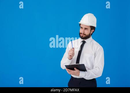 A young bearded male construction worker in a white shirt and construction helmet on a blue background. industrial engineer-Architect hold clipboard Stock Photo