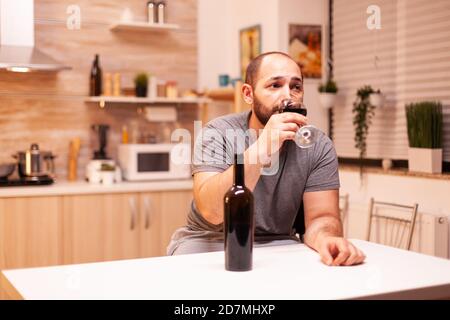 Upset young man drinking alone a bottle of red wine sitting at table in kitchen. Unhappy person disease and anxiety feeling exhausted with having alcoholism problems. Stock Photo