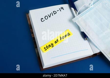 text coronavirus, stay at home, office there written on a Notepad by hand. the view from the top. mask and ballpoint pen with Notepad on a blue backgr Stock Photo