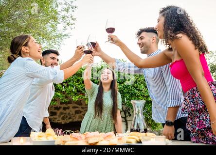 Multi ethnic happy young friends group toasting in the garden at sunset with red wine glasses - Millennials having fun together in countryside celebra Stock Photo