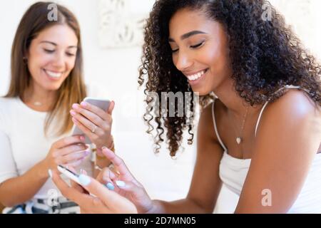 Two young beautiful women at home using smartphone - Cute black Hispanic brunette having fun indoor chatting on social networks with her beautiful blo Stock Photo